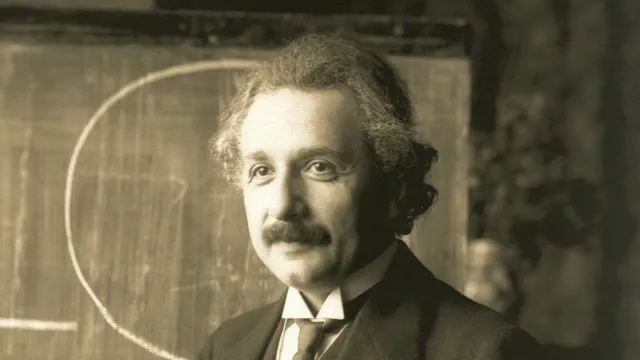 Albert Einstein in a dark suit in front of a blackboard with a chalk curve drawn on it
